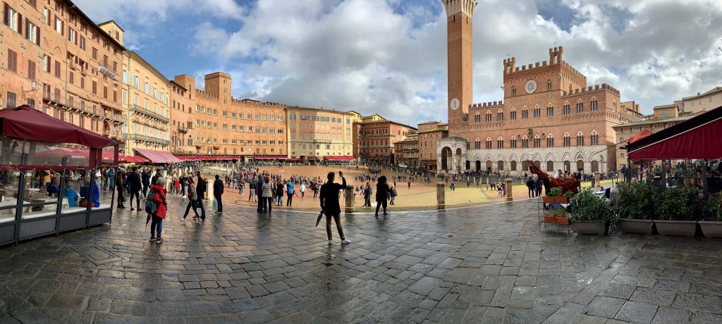 European Travelog 20191103           Siena: A perfect lunch in a perfect little town  After experiencing firsthand the effects of over tourism in Florence (I am well aware that for being there, I am contributing to the problem), Siena was a welcome respite.  A mere 72 km and slightly over an hour bus ride away, I found myself getting lost in the narrow lanes in one moment and surprised by the majestic vista of a cityscape that lined with red-roofed houses in another.    In the centre of the old city is the inviting Piazza del Campo. This is also the site for the fabled Palio horse race that occurs twice a year.  Ten horses randomly selected from the seventeen Contrade (neighbourhoods) raced around a temporary oval track, on the inner rim of the Piazza del Campo to vie for the trophy: a Palio or banner bearing an image of the Virgin Mary.  On the day of my visit, the ground still soaked with the morning rain but the sun was fighting to make an appearance.  Standing in the centre of the piazza, I could imagine the galloping of horses,  the roaring of the crowd, and the cheering of the winner.  While I was in Siena, I chanced upon Consorzio Agrario di Siena, a local supermarket with a tiny food counter that served up paninis and pizzas hot off the oven.  Squeezing myself amongst a group of hungry patrons, I devoted two heavenly slices of pizzas.  With melting mozzarella cheese, creamy tomato sauce, some hams, and dotted with a few fresh basil leaves, they were the cheapest yet the best pizzas I had on this trip.  It was such a simple delight that I was reminded how easy we could experience the blessings of life.  After filling my stomach, I walked to the bus terminal to purchase a return ticket for Florence.  Figuring out how public transportation works in a foreign country often is a challenge.  In this case, I was directed to Area B for the bus stop.  Alas, Area B was an elongated island with several signs for bus stops.  There was a dozen of passengers waiting and there wasn’t really a queue since nobody knew which was the correct loading spot.  People were milling around when a bus pulled into one side of the island.  Just when the small group of passengers surged towards it, the bus took off and swung around to the other side of the island.  Like lemmings, the crowd hurried to the other side and boarded the bus.  Sometimes, I find such inefficiency and disorder maddening.  Yet, things always seem to work out fine at the end.  Maybe it is I who need things to be orderly and efficient that need to learn to accept the natural flow of things.  Photo albums:   Siena Part 1 of 2 Siena Part 2 of 2